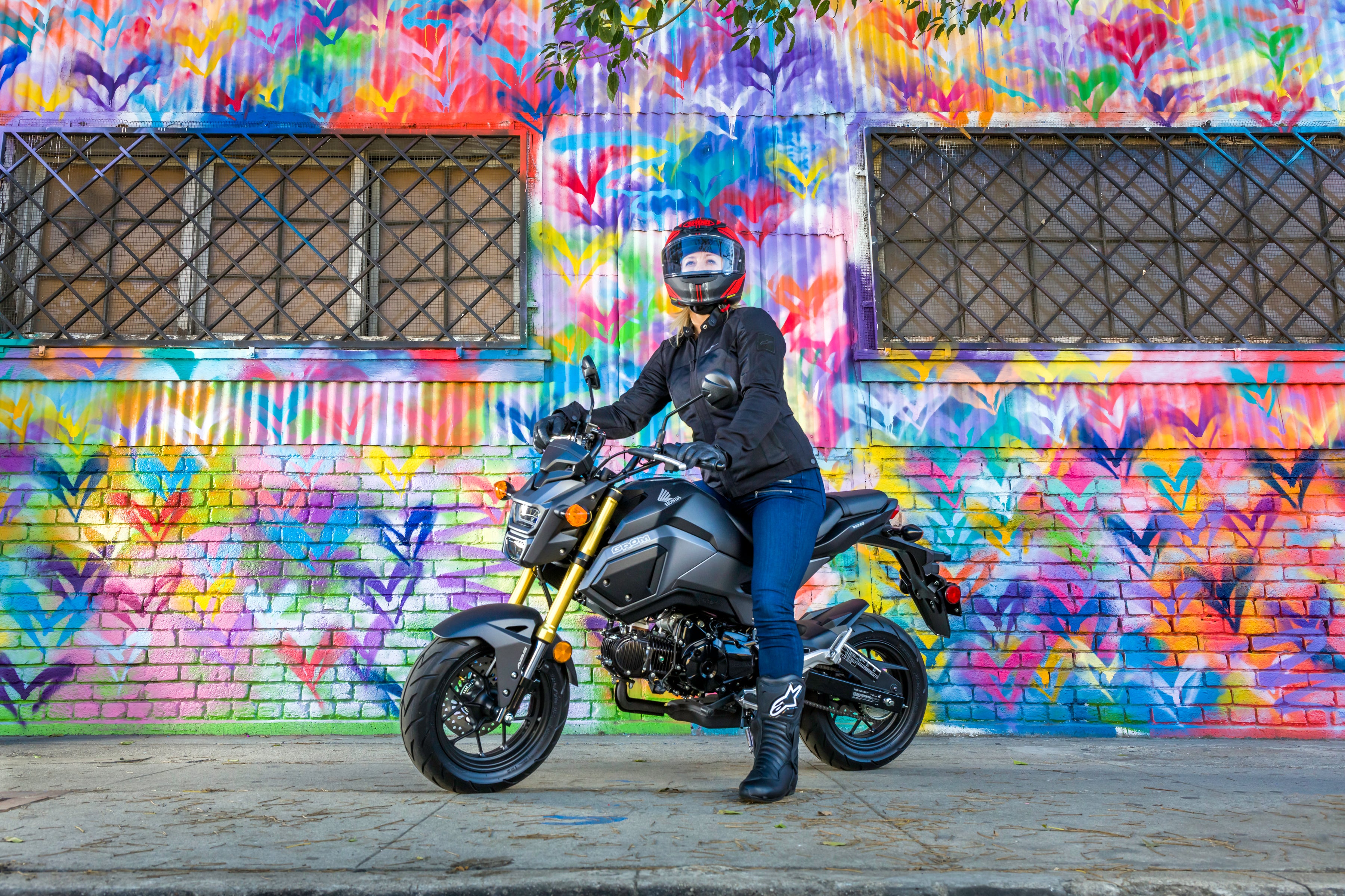 Rider in full gear posed on gray Honda Grom in front of colorful graffitied wall