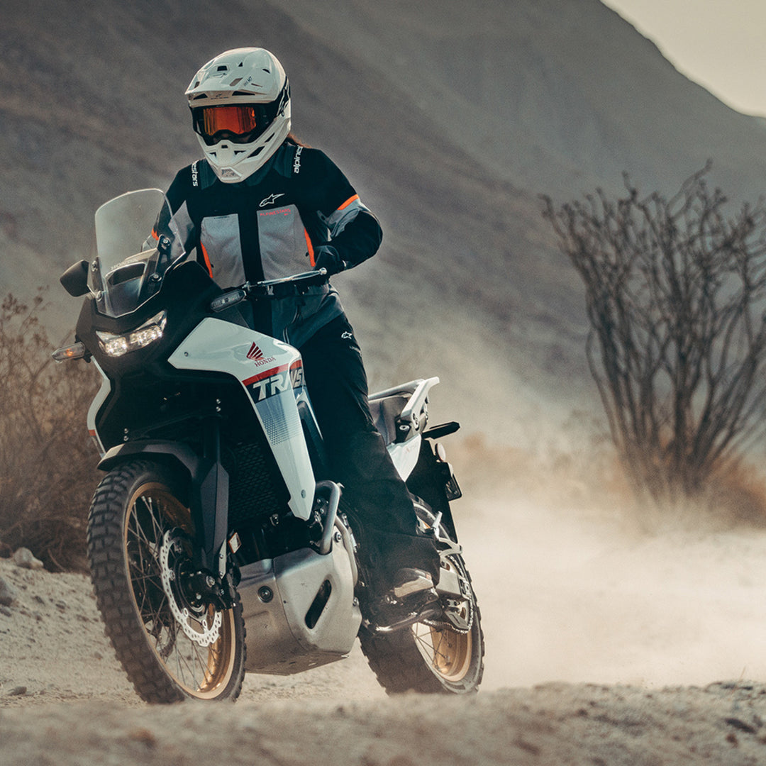 Alpinestars Stella Andes V4 Drystar Jacket Dark Gray/Ice Gray Black/Coral - Lifestyle Head-On Shot of Rider Wearing Jacket While Riding Past Mountain on White Adventure Motorcycle