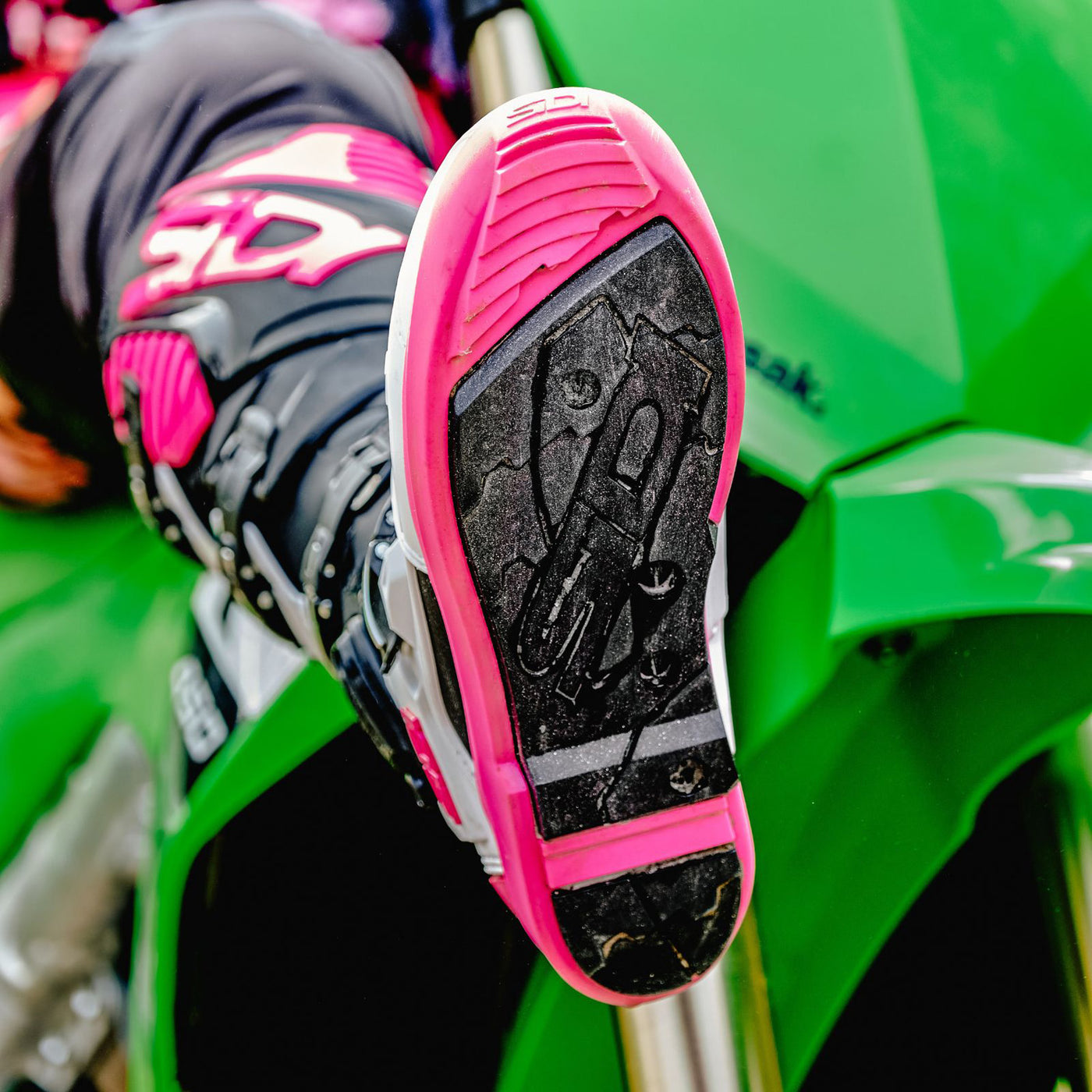 Sidi Crossfire 3 SRS Boots Black/White/Pink - Lifestyle Close-Up of Rider on Green Dirt Bike Kicking Up Leg, Holding Sole of Boot towards Camera