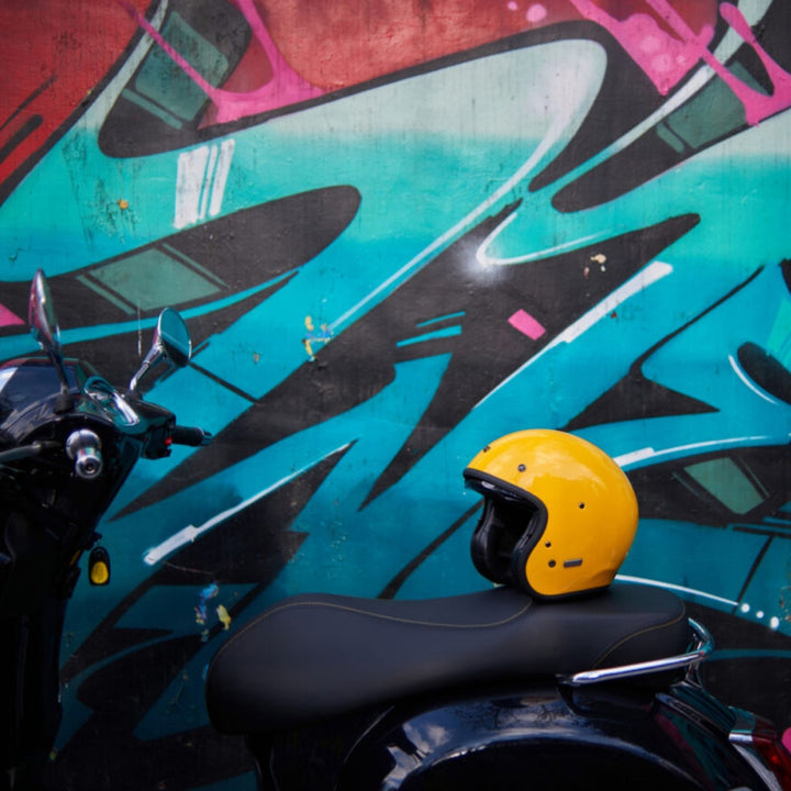 HJC V31 Solid Helmet Deep Yellow - Lifestyle Shot of Helmet Set on Scooter Seat with Graffiti Wall in Background