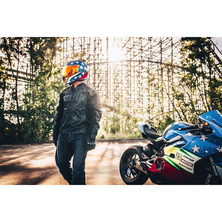 ICON Airflite™ Space Force Helmet Glory - Lifestyle Wide Shot of Rider Wearing Helmet While Standing Next to Blue Motorcycle in Front of Bridge Lattice